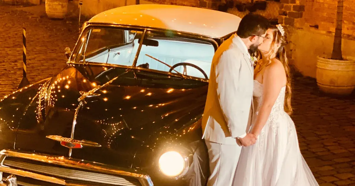 Casal se beijando ao lado de um carro antigo preto com os faróis acesos, à noite