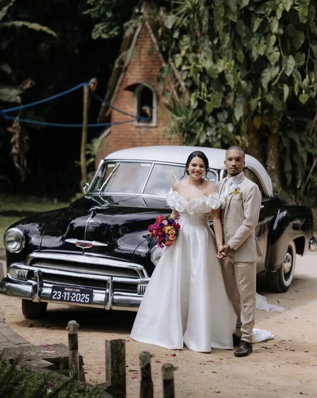 Noiva de vestido ombro a ombro e noivo de terno bege posando em frente ao Chevrolet 1951 preto
