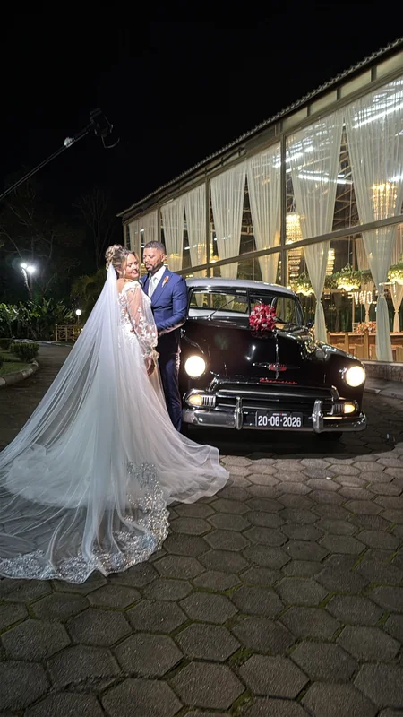 Chevrolet 1951 preto com faróis acesos à noite, noiva de véu longo ao lado e salão de festas ao fundo