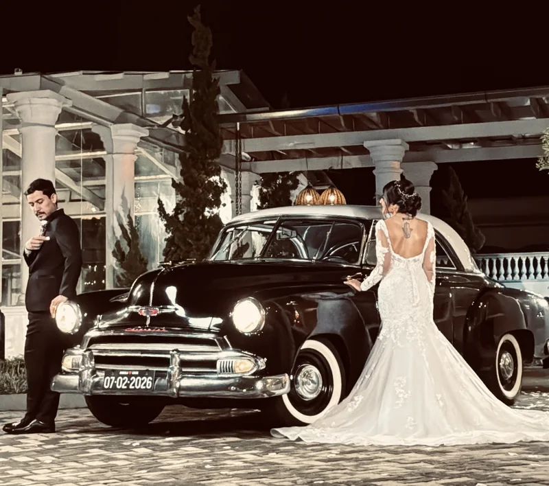 Noiva de vestido longo ao lado do Chevrolet 1951 preto com faróis acesos à noite, noivo de terno ao fundo