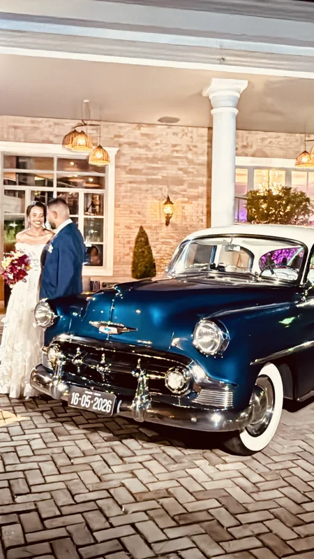 Casal ao lado do Bel Air 1953 azul em pátio de tijolinho com salão de festas ao fundo