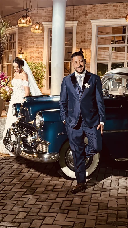 Noivo de terno azul-marinho encostado no Bel Air 1953 azul, noiva de véu ao lado