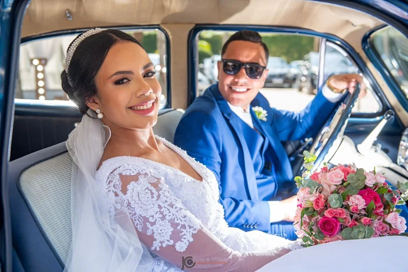 Noiva de renda e noivo de óculos escuros sorrindo no banco do Bel Air 1953, buquê rosa no colo