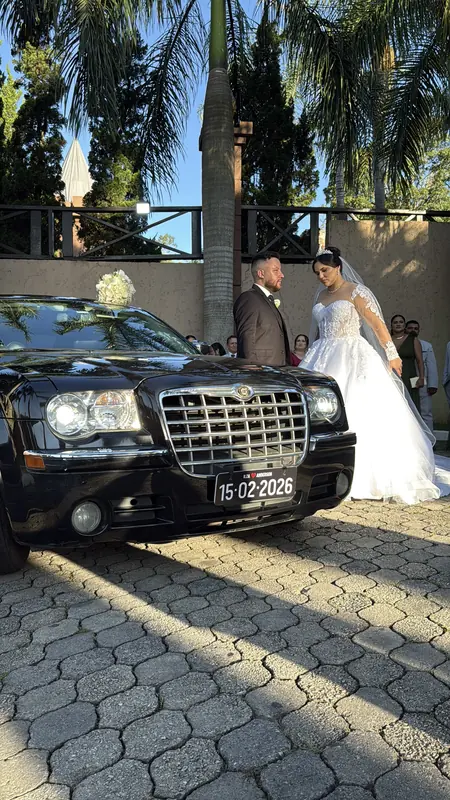 Frente do Chrysler 300C preto com grade larga e casal de noivos ao fundo, sol baixo