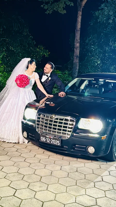 Noiva de véu e noivo apoiados no capô do Chrysler 300C preto com faróis acesos