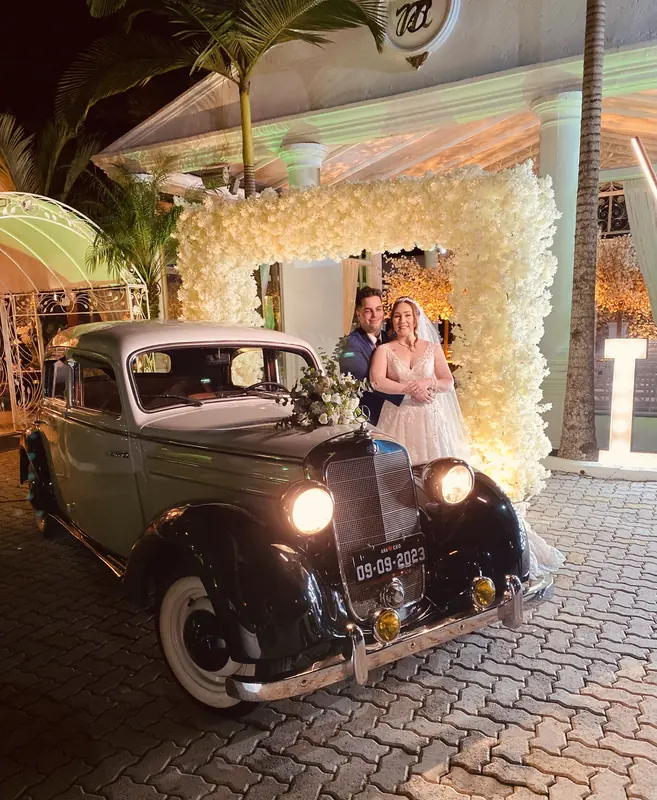 Casal de noivos atrás do Mercedes-Benz 170S em frente a arco de flores brancas e iluminação quente